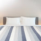 Cayambe baby alpaca wool blanket – a neatly made bed displays two white pillows resting against a gray headboard, with a striped blue and white blanket covering most of the bed in a minimal, neutral room