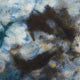 Abstract pattern featuring swirling mixes of dark brown and blue hues on a textured fabric surface, creating an impression of fluid movement and depth in an indistinct background.