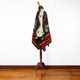 A patterned poncho with geometric designs is draped over a wooden mannequin. The setting has a wooden floor and plain white walls.