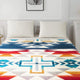 A bed features a patterned blanket with geometric designs and large crosses, beneath two white pillows. Above it hangs a framed picture labeled Plantain, in a white-walled room.