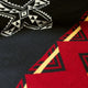 A textile showcases geometric patterns, featuring a black background with white and red contrasting triangular and diamond designs, highlighted by yellow accents, indicative of traditional or cultural motifs.