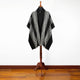 Poncho displayed on a mannequin stand, featuring a black base with white geometric patterns and buttons near the neckline. It is set against a white background on a wooden floor.