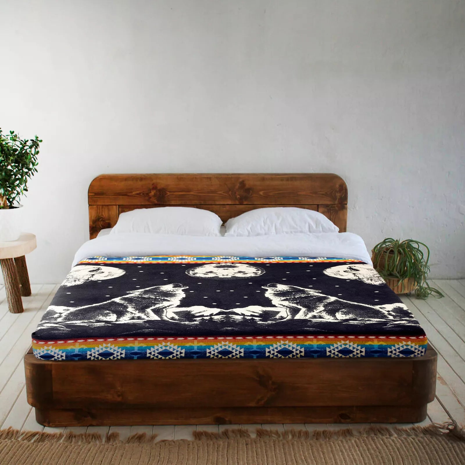 A wooden bed features a blanket depicting howling wolves under full moons, set in a minimalistic room with white walls, a potted plant on a round side table, and a wooden floor.