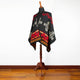 A black poncho with red geometric patterns and tan designs is displayed on a wooden mannequin stand, set against a white wall and wood floor background.