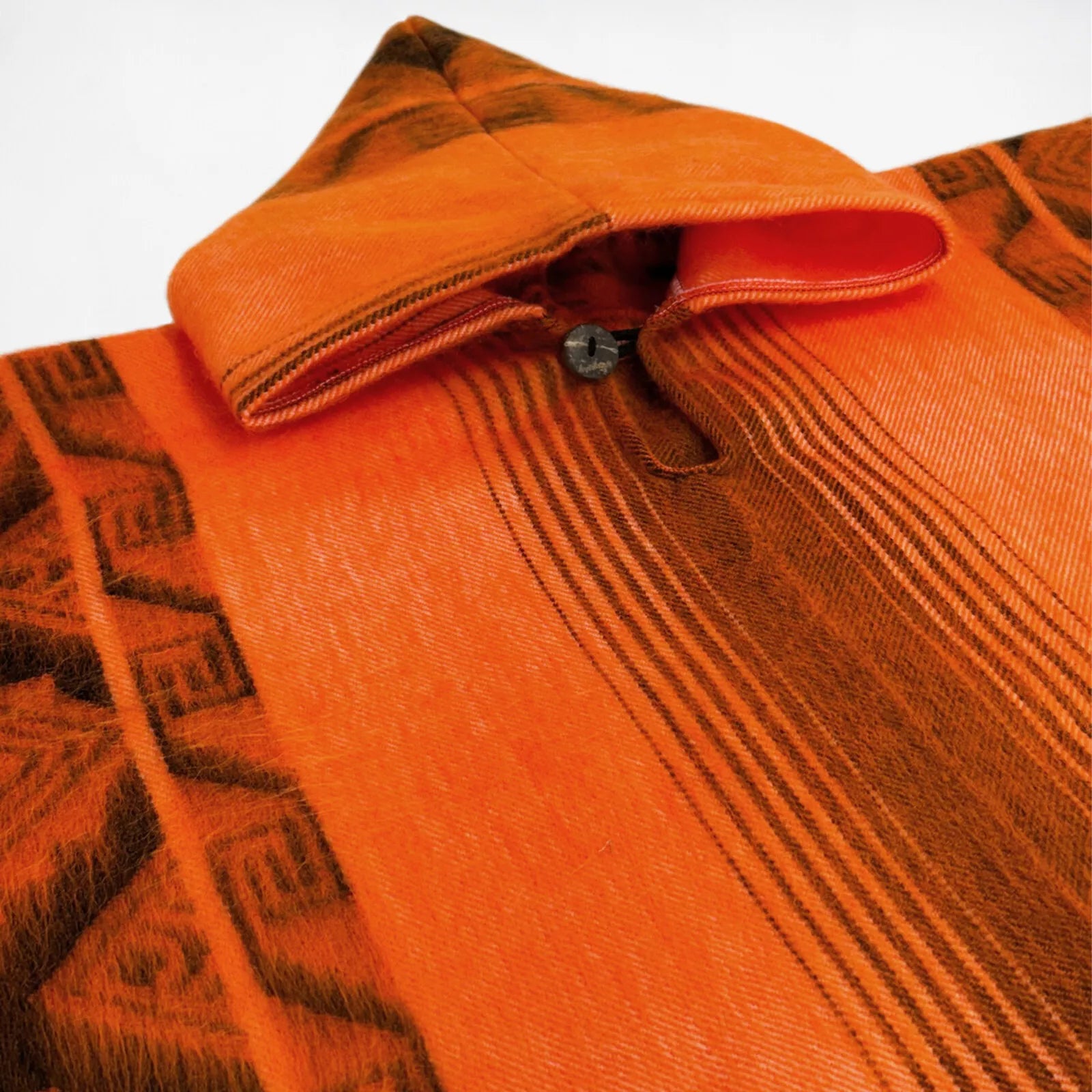 A close-up of an orange woven poncho features intricate brown geometric patterns. It includes a buttoned neckline detail, highlighting traditional textile craftsmanship against a plain background.