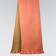 A dual-colored scarf in coral and olive drapes vertically, showcasing its soft texture and fringed ends against a plain, light-gray background.