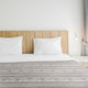 A bed with a wooden headboard is neatly made with two white pillows and a patterned grey blanket, positioned in a minimalistic room with a hanging bulb and grey curtains.
