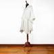 A white poncho with a hood and fringed edges is displayed on a mannequin, situated on a polished wooden floor against a plain white wall.