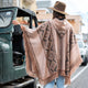 A person wearing a patterned poncho and hat leans on a green off-road vehicle, parked on a city street with buildings and cars lining the road.