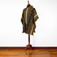 A colorful poncho with geometric patterns drapes over a wooden mannequin stand, set in a room with wooden flooring and a white wall background.