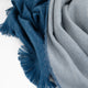 A soft, textured scarf lies crumpled, featuring intertwined blue and grey fabric with fringed edges on a plain white background.
