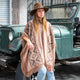 A person wearing a wide-brimmed hat and patterned poncho leans against a mud-splattered, open-top green jeep on a cobblestone street. They're gazing thoughtfully into the distance.