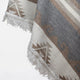 A patterned woven rug hangs with a geometric design in beige and gray. It features triangular shapes and white fringed edges, set against a neutral background.