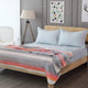 Ajilanga baby alpaca wool blanket – a neatly made wooden bed features pale blue pillows and sheets, with a striped gray and orange blanket draped over it. Nearby, a tripod lamp stands beside a wire chair, against a backdrop of gray walls adorned with geom