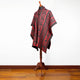 A red and black patterned poncho drapes over a wooden mannequin stand, set against a simple white wall and wooden floor.