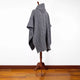 A gray knitted poncho with a high collar draped over a wooden mannequin stand, set against a white wall and wooden floor background.