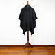 A black poncho with a hood and front pocket hangs on a mannequin stand in a room with a hardwood floor and plain white wall.