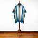 A blue and beige woven poncho with fringe hangs on a wooden mannequin stand, set against a plain white wall on a polished wooden floor.