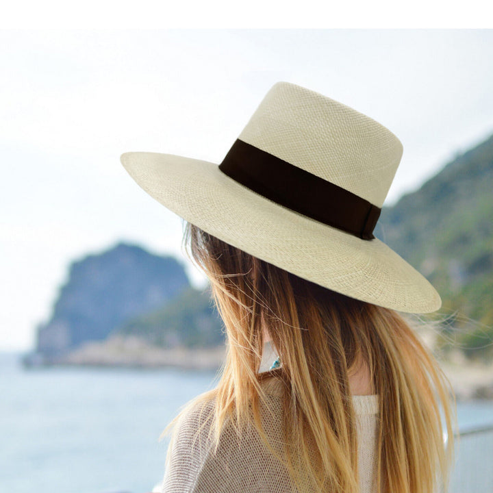 A woman wearing a wide-brimmed hat is gazing towards a mountainous coastline. The scene is by the water, with blurred greenery in the background.