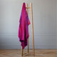 Chone baby alpaca wool blanket – a wooden clothes rack displays a vibrant pink and purple striped fabric draped over it, set against a plain light-colored wall and hardwood floor