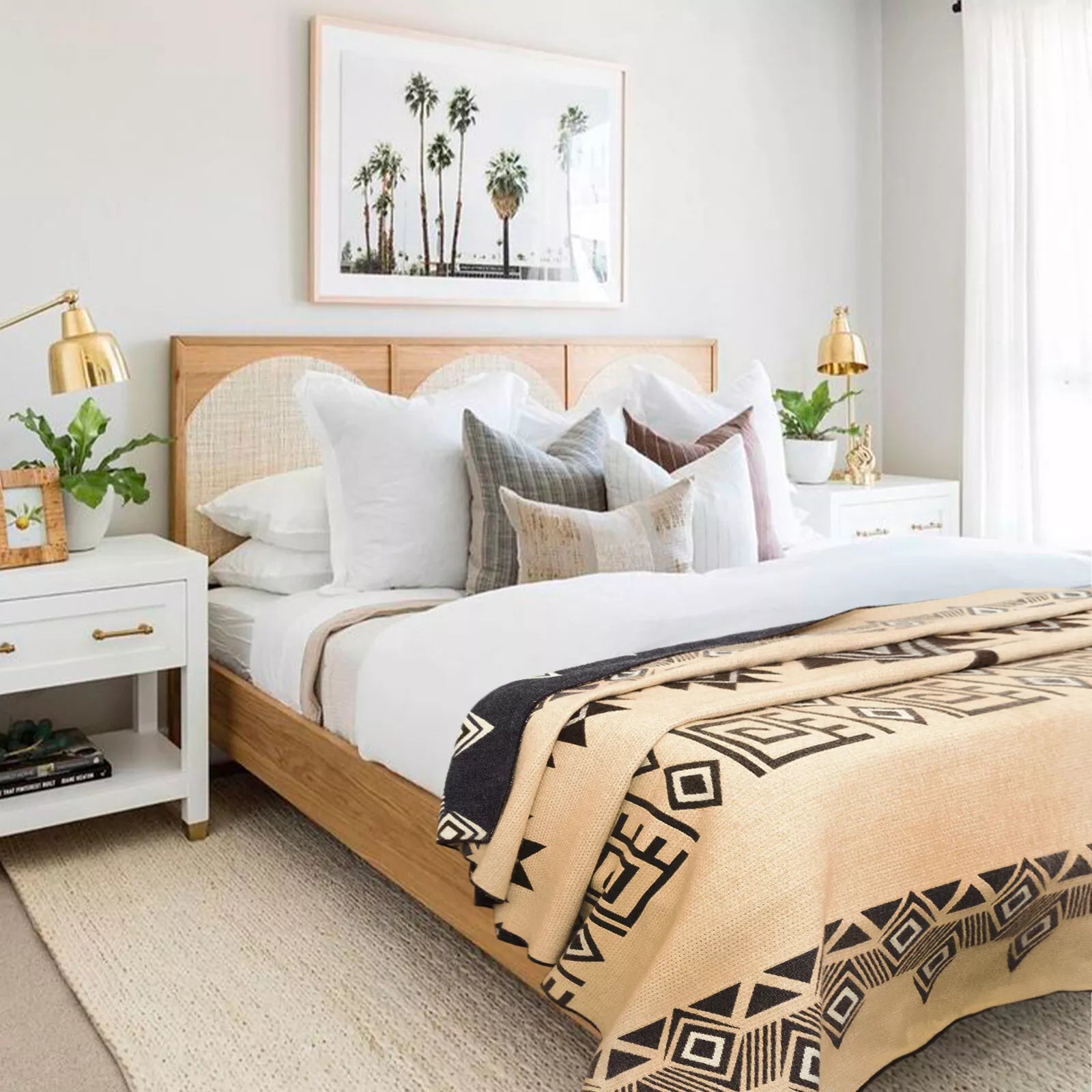 A bed, adorned with patterned blankets and numerous cushions, sits between two white nightstands. A framed palm tree picture hangs on the wall above, and plants decorate the room.