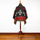 A black and red poncho with geometric patterns and crosses displayed on a wooden mannequin in a room with wooden flooring and a white wall backdrop.