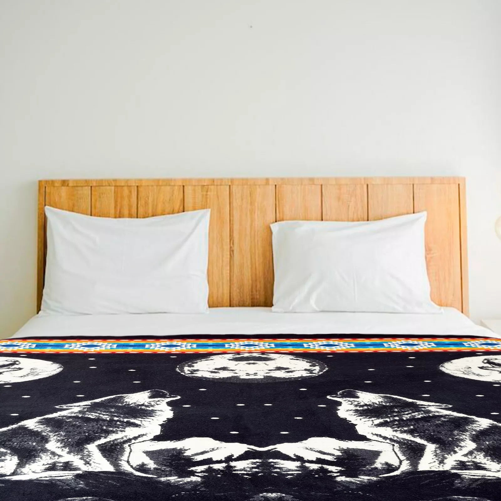 A wooden bed with white pillows and a black blanket featuring howling wolves under moons, set in a simple room with a plain white wall.