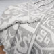 A gray and white patterned blanket lies folded on a light surface. The design features geometric shapes and abstract figures, with fringed edges enhancing its textured appearance.