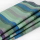 Imbabura baby alpaca wool blanket – a neatly folded blanket features wavy stripes in shades of green, blue, and purple, resting on a flat, white surface