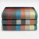 Anzu baby alpaca wool blanket – three folded blankets display a multi-colored, checkered pattern of gray, blue, orange, yellow, and red hues, stacked neatly against a plain white background