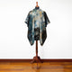 A tie-dye poncho with a hood is displayed on a wooden stand. The garment has a pattern of blues and earthy tones, situated against a plain white wall on a wooden floor.