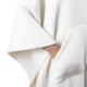 A hand is tucked into a front pocket of a soft, textured, white garment, possibly a sweater or poncho, against a plain background.
