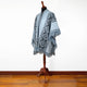 Poncho on a wooden mannequin, featuring a light blue base with intricate, dark geometric patterns and fringed edges, placed in a room with a wooden floor and plain white wall.
