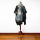 A multicolored, tie-dye cloak drapes on a wooden mannequin stand, positioned on polished hardwood flooring against a plain white wall.