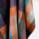 Anzu baby alpaca wool blanket – a multicolored, plaid fabric with soft hues of orange, purple, teal, and gray, hangs gracefully, showing a blend of textures in an indoor setting with subtle lighting