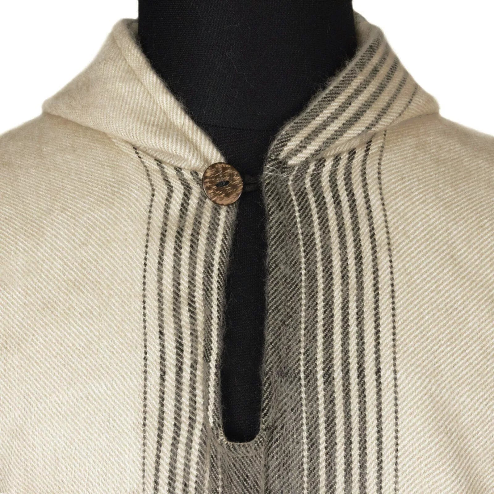 A cream-colored fabric with vertical black stripes features a hood and a brown button at the collar, displayed on a black mannequin background.