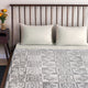 A bed with a geometric-patterned blanket and two white pillows rests against a slatted wooden headboard. A small table with orange flowers is beside the bed in a simple bedroom setting.