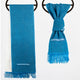 Two teal scarves with fringe hang on black rings; one drapes straight while the other is knotted. Both feature a geometric blue and white embroidered pattern.