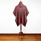A patterned red and black poncho, featuring geometric designs, drapes on a wooden stand against a plain white background and wooden floor.