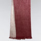 A woven scarf hangs vertically, featuring a red front and a lighter back with fringe on the bottom, set against a plain gray background.
