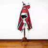 A red and green patterned poncho with geometric designs hangs on a wooden mannequin against a white wall and wooden floor, suggesting a display in a showroom or store.