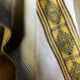 A fabric displays a folded, intricate pattern with yellow and brown tones. It features a central geometric design on a bright background, creating a textured, artistic appearance.