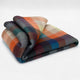 Anzu baby alpaca wool blanket – a folded, multicolored plaid blanket featuring orange, blue, and green hues rests on a smooth, light gray surface, creating a cozy and vibrant appearance