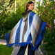 A person wears a blue-striped poncho with fringe, turning slightly to the side, in a lush garden setting with green foliage and a pathway.