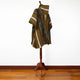 A patterned poncho with green, orange, and white stripes and geometric designs is draped over a wooden stand on a polished wooden floor against a plain white wall.