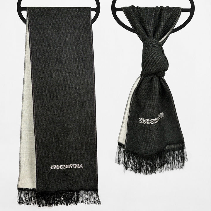 Two black scarves hang on circular holders against a plain background. One is unfolded, and the other is knotted. Both have fringe ends and embroidered white geometric patterns.