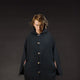 A person stands, wearing a dark, hooded cloak with buttons, staring intently. The background is a plain, dark setting, emphasizing the figure's focused expression and attire.