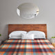 Anzu baby alpaca wool blanket – a wooden headboard frames a bed covered with a colorful checkered blanket. Above, an ornate oval mirror reflects part of the room, highlighting a minimalist decor with plants