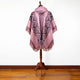A pink and black patterned poncho is displayed on a mannequin with wooden floors and a plain white background. The poncho features a hood and fringe at the edges.