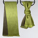 Two green fringed scarves hang on black circular hooks against a light gray background. One scarf drapes straight, while the other is tied in a knot.